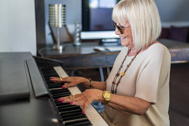 Smiling senior woman wearing sunglasses playing piano while sitting at home - DLTSF01037