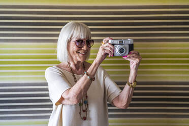 Lächelnde ältere Frau mit Sonnenbrille beim Fotografieren mit der Kamera an der Wand zu Hause - DLTSF01048