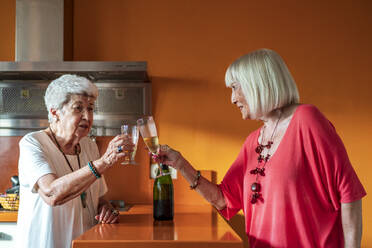 Senior female friends toasting champagne while standing in kitchen at home - DLTSF01052