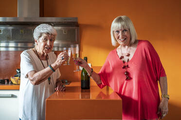 Smiling senior female friends toasting champagne flutes while standing at home - DLTSF01053