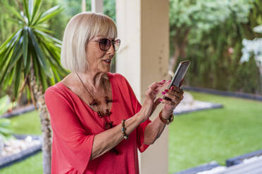 Stilvolle ältere Frau mit Sonnenbrille, die ein Mobiltelefon benutzt, während sie auf der Terrasse steht - DLTSF01056