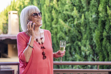 Senior woman holding champagne flute talking over mobile phone while standing in balcony - DLTSF01058