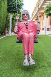 Stilvolle ältere Frau mit Sonnenbrille auf einer Schaukel im Garten sitzend - DLTSF01063