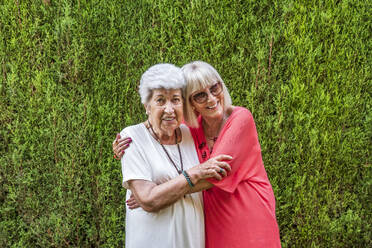 Smiling senior female friends embracing while standing against plants in yard - DLTSF01065