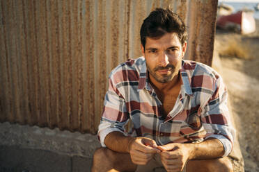 Close-up of confident man sitting against wall at sunset - MPPF01004