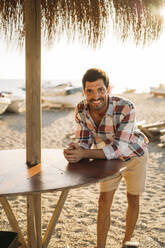 Happy mid adult man standing by table at beach during sunset - MPPF01005