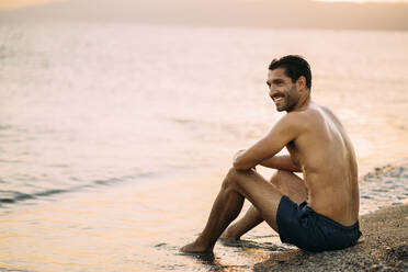 Hemdloser gut aussehender Mann, der bei Sonnenuntergang am Ufer sitzt und wegschaut - MPPF01011