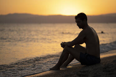 Shirtless thoughtful mid adult man sitting at shore against sky during sunset - MPPF01012