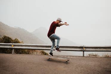 Seitenansicht eines jungen Mannes, der auf einem Longboard auf einer abgelegenen Bergstraße fährt und vor einer nebligen Landschaft springt - ADSF09555