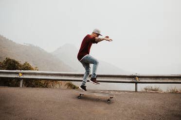 Side view of young man riding long board on remote mountain road and jumping against foggy landscape - ADSF09555