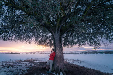 Seitenansicht eines Mannes mittleren Alters in warmer Kleidung, der unter einem Baum auf einem verschneiten Landschaftshintergrund Gitarre spielt - ADSF09588