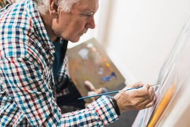 Seitenansicht eines älteren männlichen Künstlers in Freizeitkleidung, der ein Bild mit Pinsel auf Papier auf einer Staffelei malt und eine Palette mit Farben in einer Kunstwerkstatt hält - ADSF09591