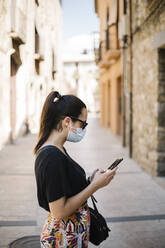 Woman wearing protective ask and using smartphone in alley - XLGF00442