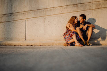 Man and woman looking at each other and embracing sitting at nearby street wall - ADSF09652