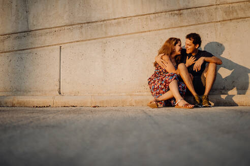 Mann und Frau schauen sich an und umarmen sich, während sie an einer nahegelegenen Straßenmauer sitzen - ADSF09652