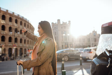 Seitenansicht einer asiatischen Frau mit Koffer, die lächelt und den Arm ausstreckt, während sie auf der Straße einer Touristenstadt ein Taxi ruft - ADSF09654