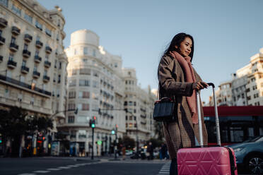 Full body ethnic woman with suitcase standing on pavement near road and looking away while visiting city on sunny day - ADSF09657