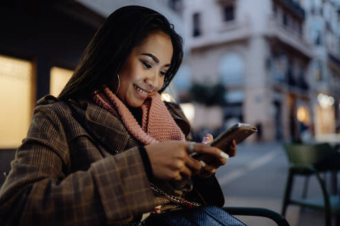 Fröhliche ethnische Frau lächelt und surft in den sozialen Medien auf ihrem Smartphone, während sie sich abends in einem Straßenrestaurant ausruht - ADSF09660