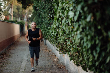 Smiling active mature man in good sportive shape jogging in narrow street along houses and botanical fence in summer day - ADSF09661