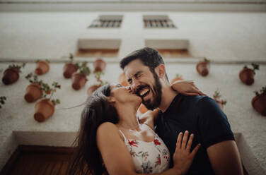 Low angle of loving casual couple having fun and cuddling while standing next to picturesque white building in summer town - ADSF09667