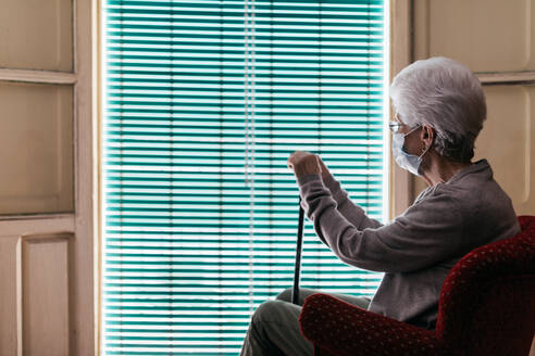 Elderly woman in protective mask and gloves with walking stick sitting near window - ADSF09692