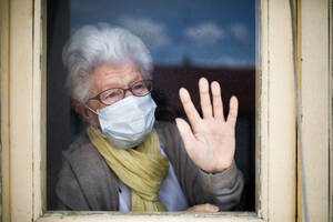 Senior woman in protective mask looking out window - ADSF09702