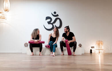 Young man and women with mats speaking with each other while sitting in white block in spacious studio during yoga training - ADSF09706