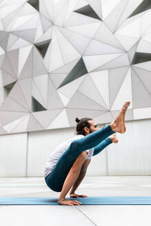 Seitenansicht eines erwachsenen bärtigen Mannes, der eine Yoga-Glühwürmchen-Pose gegen eine Wand mit geometrischem Ornament einnimmt - ADSF09743