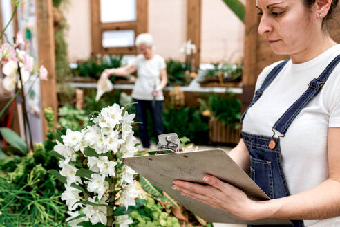 Crop adult woman writing on clipboard while standing near plant with white flowers during work in hothouse - ADSF09756