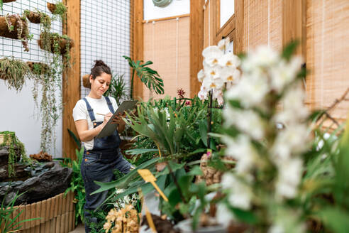 Seitenansicht einer erwachsenen Frau, die auf einem Klemmbrett schreibt, während sie in der Nähe einer Pflanze mit weißen Blüten bei der Arbeit im Gewächshaus steht - ADSF09757