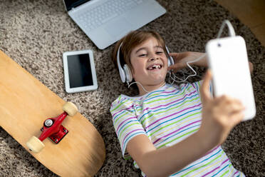 From above of cheerful little boy in casual shirt listening to music with headphones while lying on carpet near gadgets and skateboard in bedroom - ADSF09760