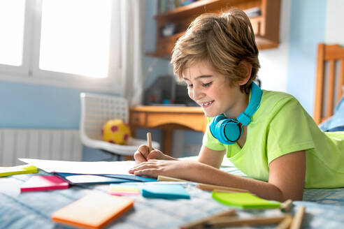 Side view of positive schoolboy in casual wear and wireless headphones enjoying music and drawing with pencils while spending free time in bedroom - ADSF09764