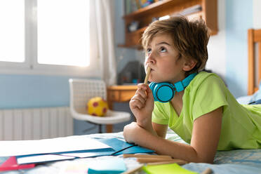 Thoughtful schoolboy in casual wear and wireless headphones enjoying music and drawing with pencils while spending free time in bedroom - ADSF09765