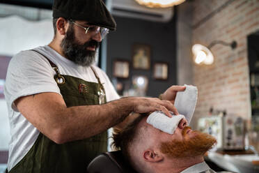 Barbier macht Gesichtsmassage zu gut aussehend rothaarigen Mann mit geschlossenen Augen sitzen in Stuhl mit warmen heißen Handtuch bedeckt Gesicht - ADSF09780