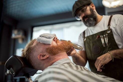 Barbier mit Kamm und Trimmer Schneiden Rothaarige Mann Bart mit Handtuch bedeckt Augen sitzen in Barbershop - ADSF09781