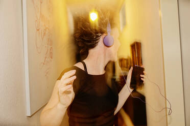 Carefree mid adult woman with headphones dancing at illuminated home - JATF01215