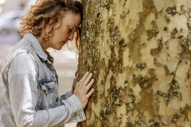 Traurige erwachsene Frau mit geschlossenen Augen, die sich an einen Baumstamm im Park lehnt - JATF01228