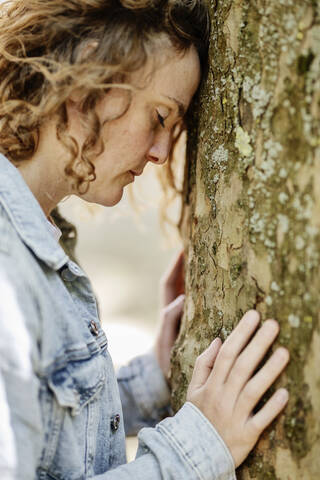 Close-up of sad woman with eyes closed leaning on tree trunk in park stock photo