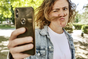 Mid adult woman making face while taking selfie with mobile phone in park - JATF01235