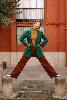 Young woman wearing green jacket standing on bollards against building in city - TCEF00972