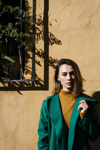 Schöne Frau mit grüner Jacke steht an einer Wand an einem sonnigen Tag, lizenzfreies Stockfoto