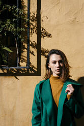 Beautiful woman wearing green jacket standing against wall on sunny day - TCEF00974