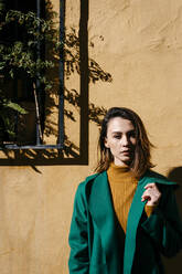Schöne Frau mit grüner Jacke steht an einer Wand an einem sonnigen Tag - TCEF00974