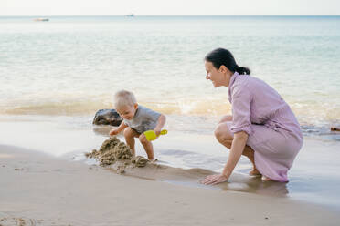 Kleinkind und Mutter spielen mit Sand am Strand gegen unscharfe Meereslandschaft in sonnigen Tag - ADSF09830