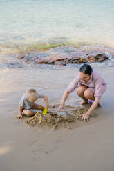 Toddler and mother playing with sand on beach against blurred seascape in sunny day - ADSF09831