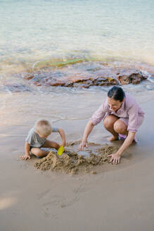 Kleinkind und Mutter spielen mit Sand am Strand gegen unscharfe Meereslandschaft in sonnigen Tag - ADSF09831