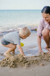 Kleinkind und Mutter spielen mit Sand am Strand gegen unscharfe Meereslandschaft in sonnigen Tag - ADSF09832