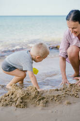 Toddler and mother playing with sand on beach against blurred seascape in sunny day - ADSF09832