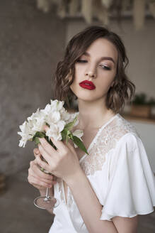 Charmante bescheidene Dame im weißen Kleid mit Spitze schaut weg und träumt, während sie ein Glas mit kleinen weißen Blumen hält und den Duft genießt - ADSF09839