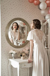 Attraktive ernsthafte junge Frau steht und schaut in großen runden Spiegel im Boudoir - ADSF09842
